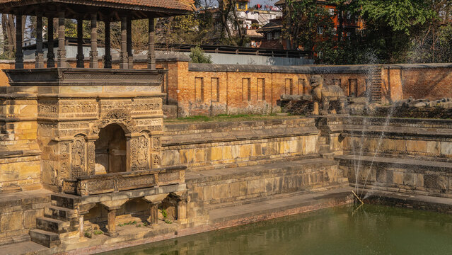 Ancient Newar architecture. A pool with stone terraces and steps. Exquisite carvings on the columns and walls of the gazebo. Fountain spray. Sculpture of a dog on the shore. Nepal. Patan.