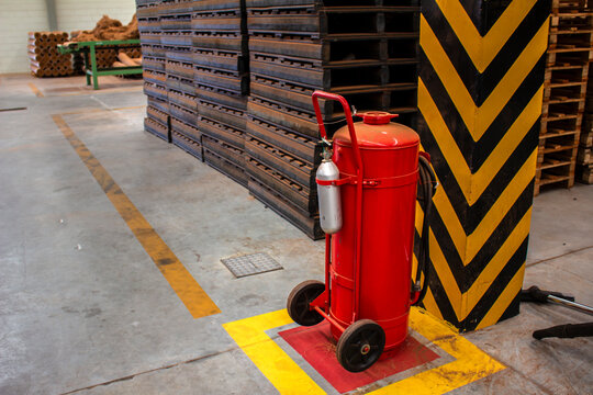 A large mobile red fire extinguisher on wheels positioned in an industrial warehouse setting with safety markings on the floor, providing emergency fire protection and workplace safety in Brazil.