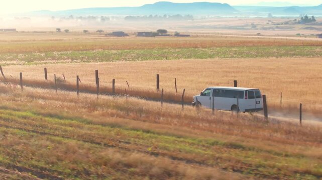 White passenger van travels along dusty dirt road through expansive farmland with golden wheat fields and green crops. Mountains visible in misty morning light with fence posts lining the countryside.