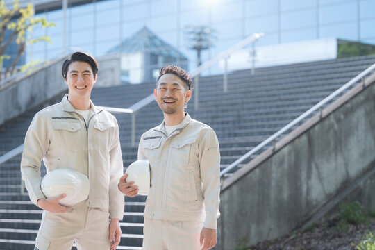 ヘルメットを持つ若手作業員のポートレート メンテナンスやゼネコンのような建設現場と成長、ビル背景でカメラ目線の作業着の2人　右にコピースペースあり