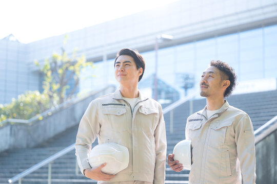 ヘルメットを持つ作業員のポートレート メンテナンスやゼネコンのような建設現場と成長、ビル背景で見上げる作業着の2人