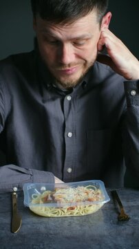 Melancholic man sitting at dark table receives prepackaged pasta meal in plastic container, expressing sadness, disappointment about instant lunch delivery service. Vertical shot.
