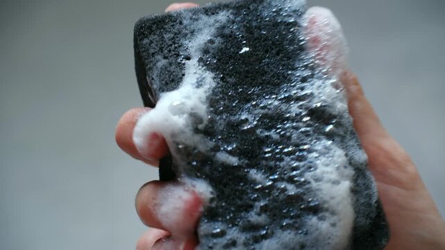 Close up human hand holding, repeatedly squeezing black cleaning sponge, creating white soapy foam bubbles that drip down, showing cleaning or washing concept. Hand squeezing black sponge with foam