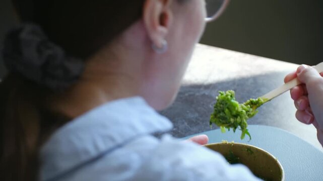 Over shoulder view businesswoman in glasses eating fresh vegetable salad with wooden fork from paper bowl during healthy lunch break at office. Woman eating healthy green salad bowl for lunch