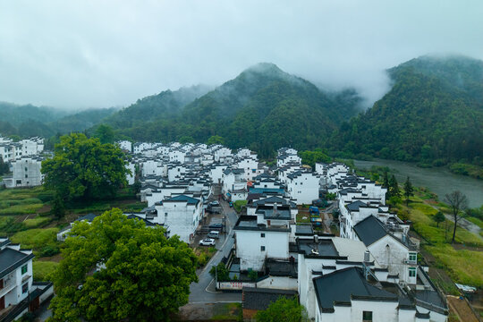 An aerial view shows the traditional white houses of Huizhou Ancient City located in She County Anhui province China surrounded by green misty mountains and a calm river under a cloudy sky