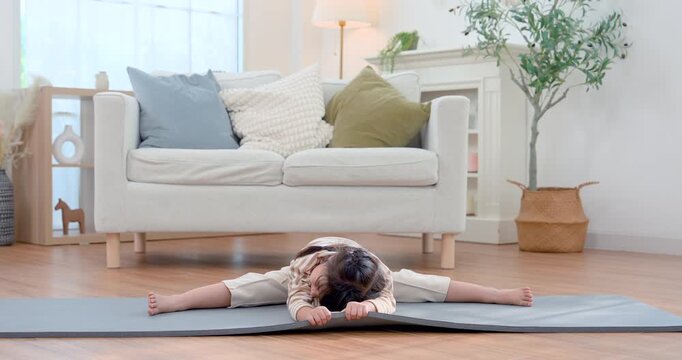Asian preschool girl leans forward in wide leg stretch on exercise mat in bright living room, practicing yoga with steady movement and body control, building flexibility, balance, strength, wellness.