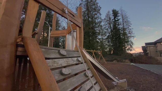 Childrens play equipment featuring rope pull climbing wall constructed from natural timber standing in neighborhood park under vibrant orange cloudy sunset sky captured via lateral camera sweep