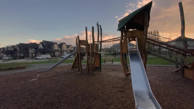 Childrens play equipment featuring rope pull climbing wall constructed from natural timber standing in neighborhood park under vibrant orange cloudy sunset sky captured via lateral camera sweep