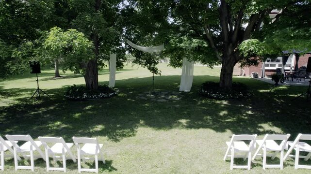 A smooth drone shot pulling back from a draped fabric wedding arch between two large trees to reveal rows of white folding chairs on a lush green lawn, perfect for wedding and event themes.