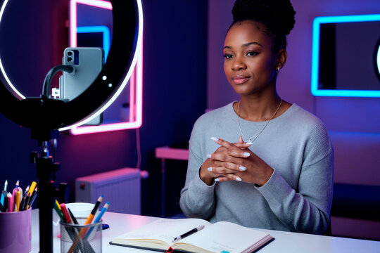 Portrait of young Black woman sitting at desk recording video with smartphone mounted on ring light, hands clasped, looking into camera, open notebook and pens on table