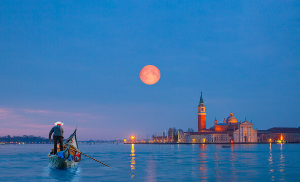 View of San Giorgio Island in Venice with wooden buoys in Giudecca Canal with full moon - Venetian gondolier punting gondola through canal waters at night - Venice Italy