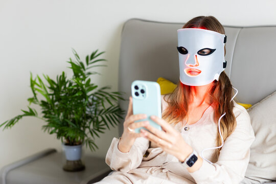 Woman using LED facial mask while checking smartphone at home