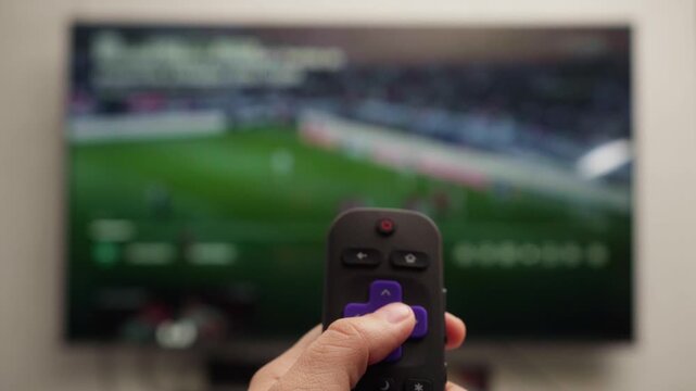 Close-up of a hand holding a remote control and pressing the buttons to navigate the menu on a smart tv, with a soccer game playing on the out-of-focus background screen
