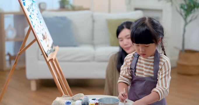 Young Asian mother watches preschool girl lift brush toward easel canvas in warm home, supporting drawing practice and creative exploration through gentle guidance, patience, bonding, early education.