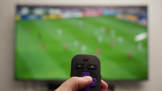 Person changing channels with a roku remote control while watching a live soccer game on a smart television, relaxing in a living room and enjoying free time at home