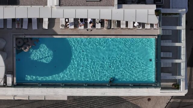 Aerial view of a luxury swimming pool from a top-down perspective with people relaxing on sun loungers and clear blue water.