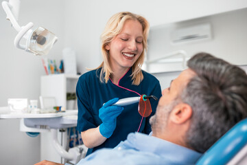 Fototapeta premium Female dentist using dental curing light on male patient. Professional teeth whitening, filling procedure, and healthcare concept. Woman doctor performing treatment in modern stomatology clinic room.