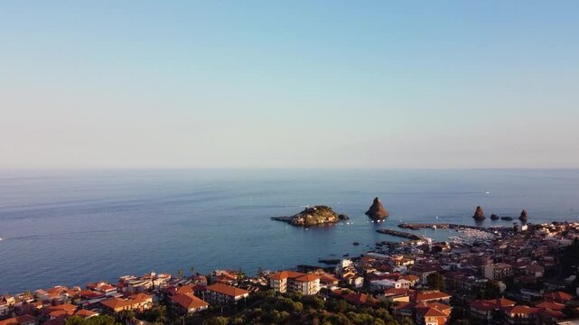 High aerial drone shot over Aci Trezza, Sicily. The historic town and harbor in the foreground, with the iconic Faraglioni dei Ciclopi sea stacks rising from the calm Mediterranean under a clear blue