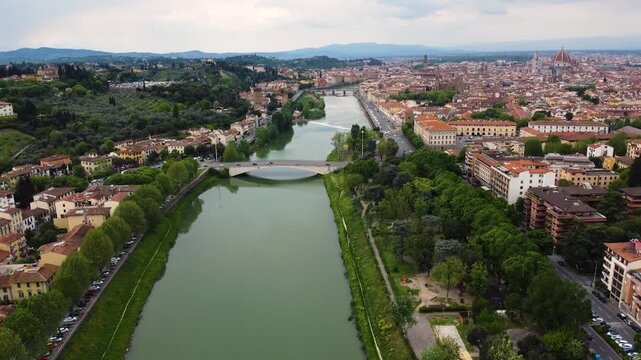 Aerial drone shot over the Arno river flowing through Florence, Italy. Historic city skyline with the Cathedral of Santa Maria del Fiore dome visible in the background under a cloudy sky.