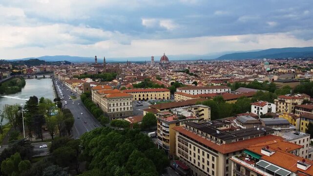Aerial drone panorama over Florence, Italy. The Arno river runs along the left, with the historic city center, Cathedral dome and Giotto's bell tower dominating the skyline under a cloudy sky.