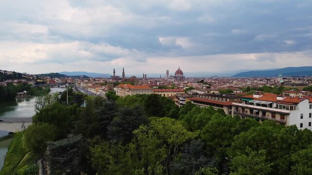 Drone panorama over Florence, Italy. Dense tree canopy in the foreground frames the full historic skyline with the Cathedral dome, Palazzo Vecchio tower and Tuscan hills under a dramatic cloudy sky.
