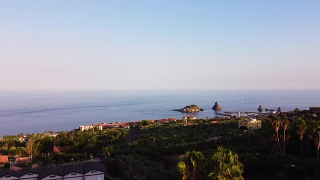 Drone shot slowly pushing toward the Faraglioni dei Ciclopi from above Aci Trezza, Sicily. Palm trees and rooftops in the foreground, the iconic sea stacks and harbor gradually approaching across the