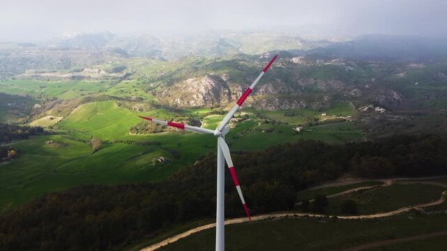 Aerial drone shot of a wind turbine at the Parco Eolico di Mineo, Sicily. Rolling green hills, rocky outcrops and the Sicilian interior landscape stretch into the distance behind the rotating blades.