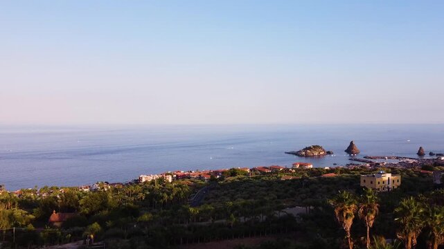 Wide aerial drone shot over Aci Trezza, Sicily. Lush Mediterranean vegetation and rooftops in the foreground, the Faraglioni dei Ciclopi sea stacks and harbor visible along the calm coastline at dusk.