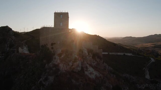 Drone shot of the medieval Castello di Serravalle perched on a rocky hilltop in Sicily, sun setting dramatically behind the ancient stone tower. Wind turbines and rolling Sicilian hills visible in the