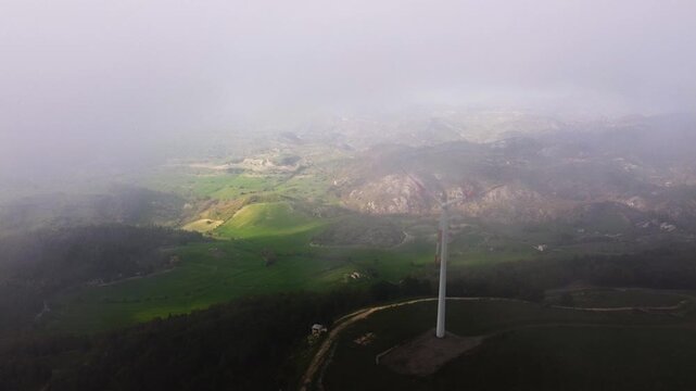 Aerial drone shot through low clouds above a wind turbine at the Parco Eolico di Mineo, Sicily. Misty atmosphere partially veils the rolling green Sicilian hills and rocky outcrops below.