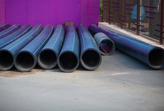 Stack of new black HDPE plastic pipes with blue stripes for cold water supply. Industrial construction site and municipal infrastructure background
