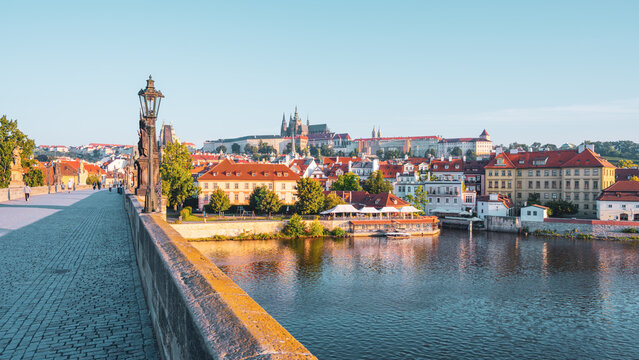 A stunning view showcases the historic Charles Bridge leading to Prague Castle, featuring cobblestone pathways and charming rooftops under a clear blue sky.