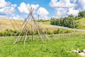 Wooden frame of conical hut reconstruction on green meadow with hills and road in Kemerovo Oblast Kuzbass © Dmitrii
