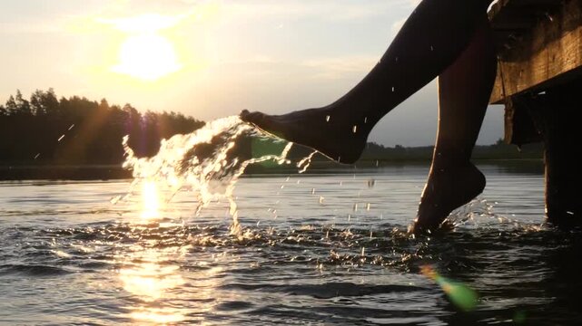 Silhouette of a person's legs dangling from a wooden pier, kicking and splashing water in a calm lake during a beautiful sunset, creating sparkling drops against the golden sunlight