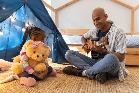Indian dad and African American daughter sitting on bedroom rug, playing guitar, she hugging teddy