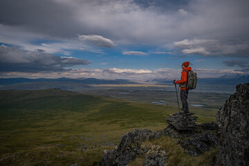 Fototapeta premium Male hiker with a backpack and trekking poles standing on a rocky peak and looking at a vast mountain valley under a dramatic sky. Traveler in an orange jacket exploring wild remote nature.