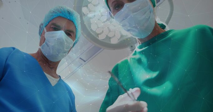 Leaning two clinicians in scrubs and masks performing surgery at OR table, holding tool under lamp