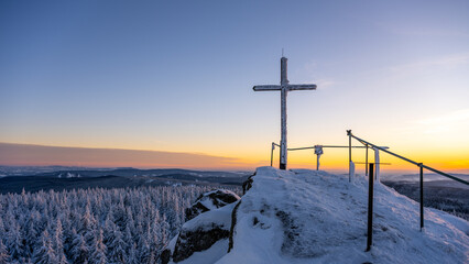 A cross stands atop the rocky summit of Jizera Mountain. Snow covers the ground and trees. The sky shows light from the setting sun as winter takes hold in the mountains. © pyty