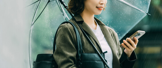 雨の日に傘を差しながらスマートフォンを使う日本人女性・ビジネスウーマンのビジネスイメージ（LP・バナー向け）, Japanese Businesswoman Using Smartphone on Rainy Day © Trickster*