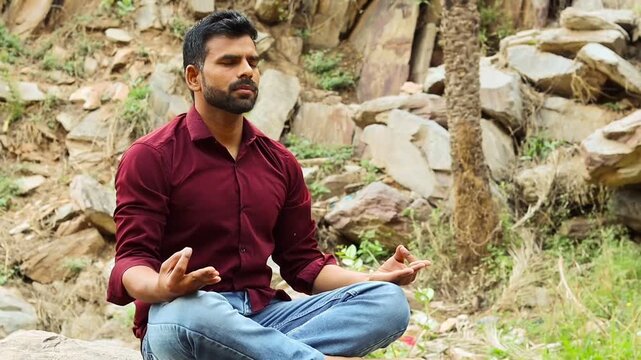 Indian man sitting in lotus pose practicing yoga and meditation on rocks, young male doing gyan mudra in nature, mental wellness and mindfulness outdoors
