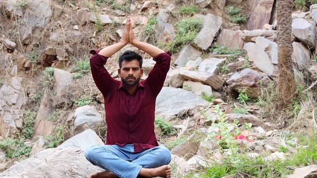 Young Indian man practicing seated yoga pose with arms raised in nature, male yogi doing namaste overhead on rocky terrain, outdoor spiritual wellness and fitness
