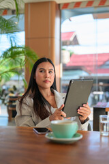 Fototapeta premium A professional Asian woman from Thai Isan looking away while holding a tablet in an authentic cafe.