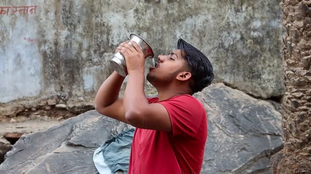 Young Indian man drinking water from traditional steel glass outdoors, thirsty male hydrating himself in summer heat, rural lifestyle and health concept
