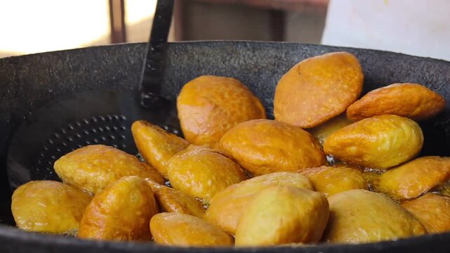 Deep frying traditional Indian kachori or puri in hot oil, close up of golden brown snacks being fried in a large iron pan, street food cooking process
