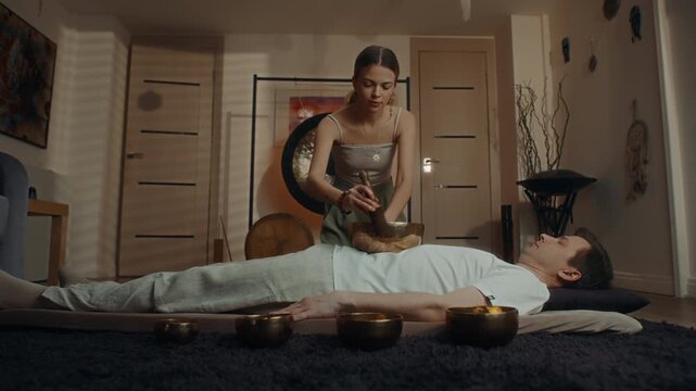 Sound healer playing singing bowls over relaxed client indoors