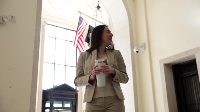 Confident young professional woman in a stylish suit walking through an elegant government hallway, talking while sipping coffee from a travel mug with american flags behind her