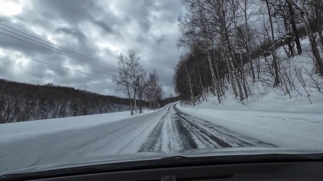 POV driving along a winding snow covered mountain road in Hokkaido, Japan, featuring curve warning signs, forest and dramatic cloudy winter sky