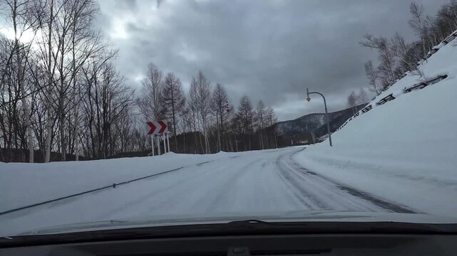 POV driving along a snow covered curved road with chevron warning signs, forest slopes and icy conditions under cloudy winter sky