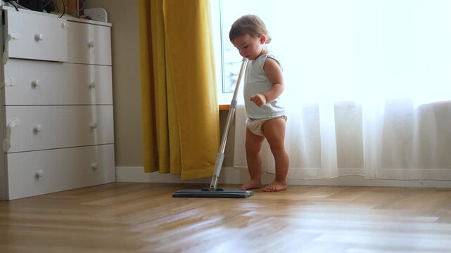 Toddler mopping hardwood floor at home. Cute baby helps with cleaning in sunny bedroom. Little child holds mop and cleans wooden floor. Toddler doing housework indoors. Baby learning to mop by window.