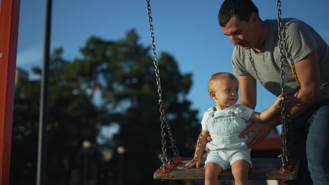 Father pushes baby on swing at outdoor playground. Dad holds toddler on chain swing with care. Baby sits on swing as father smiles nearby. Parent and child enjoy playground together under blue sky.